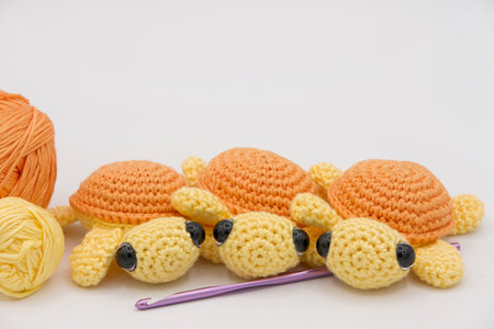 Close up on hand made crochet turtles in yellow and orange on an off white background with crochet hook and balls of yarn on the side.の写真素材