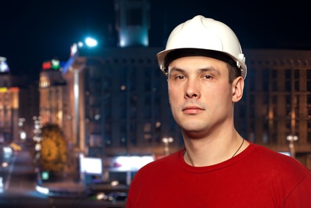 Young worker wearing a protective helmet standing against night city background.