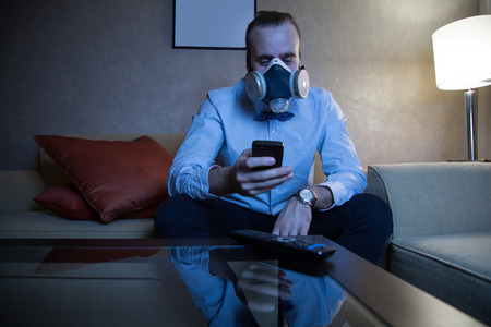Handsome young caucasian man in a blue shirt and respirator watching TV and looking at phoneの写真素材