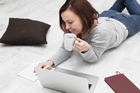 Beautiful woman with a laptop lies on the floor in the living room. Freelancer working at home indoors.の写真素材