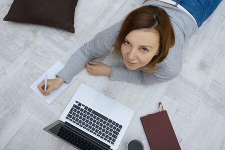 Beautiful woman with a laptop lies on the floor in the living room. Freelancer working at home indoors.の写真素材