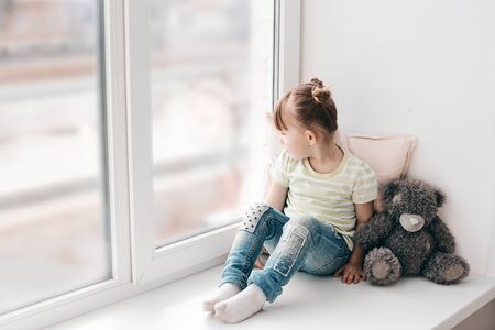 A little girl sits on a window sill and looks out the window into the street. Its a sad mood. Illness or self-isolation.の写真素材