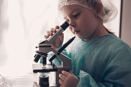 Little girl in lab coat learning chemistry in school laboratory. Young scientist in protective glasses making experiment in lab or chemical cabinet. Looking through the microscope. High quality photoの写真素材