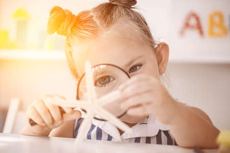 Portrait of cute adorable girl looking at shell through magnifying glass. Child with loupe studying...