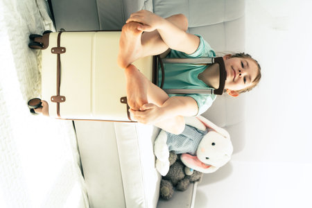 Little girl - traveler, smiling teenage girl - tourist sitting with luggage, travel suitcase, making funny face, preparing for summer travel and vacation, looking at camera, on sofa. High quality photoの写真素材