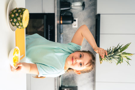 Little girl with pineapple in the kitchen. Fruit salad and pineapple are on the table. Above her head, she holds a green tuft of a pineapple. Selected focus. High quality photoの写真素材