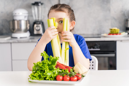 Beautiful little girl with celery in her hands and a plate of fresh vegetables. Girl in the kitchen at home on a plate of tomatoes, green salad, peppers and cheese. High quality photoの写真素材