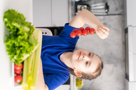 Cute little girl holding a sprig of cherry tomatoes in her hands. A girl in the background of the kitchen at the table with a plate of fresh fruit and salad. Selected focus. High quality photoの写真素材