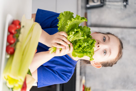 Cute little girl eating lettuce in the kitchen of the house, in front of her is a plate of fresh vegetables. Selected focus. High quality photoの写真素材