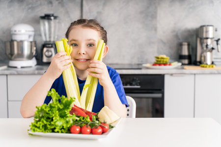 Happy, cheerful little teenage girl with celery and a plate of fresh vegetables and lettuce sitting in the kitchen at home. Proper and healthy nutrition. Selected focus. High quality photoの写真素材