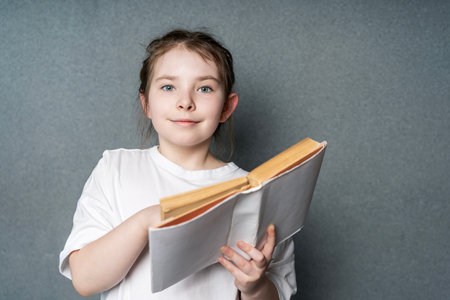 Cute little girl with an open book in her hands, looking at the frame on a gray background in the...
