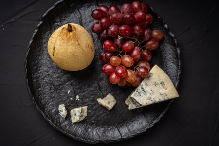 Blue blue cheese Garganzola or Dor Blue with a bunch of red grapes and a yellow pear on a black mica board on a dark background. Space for text. Top view. High quality photoの写真素材