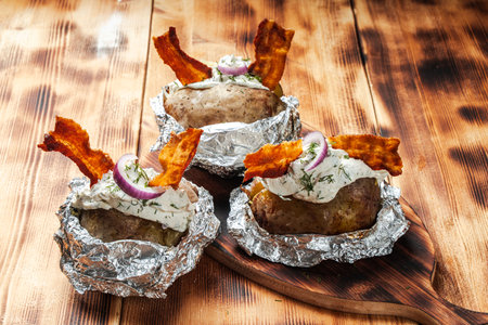 Potatoes baked in foil with cheese sauce and parsley, with onions and fried bacon on a wooden background. High quality photoの写真素材