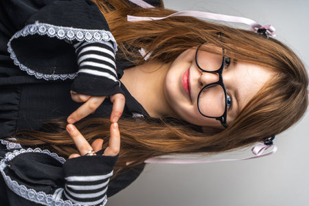 Cute little teenage girl in a black dress, mittens and glasses showing a heart on a gray background with her hands. High quality photoの写真素材