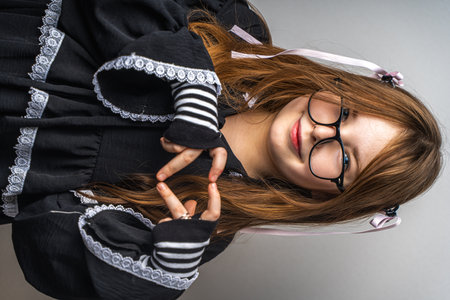 Cute little teenage girl in a black dress, mittens and glasses showing a heart on a gray background with her hands. High quality photoの写真素材
