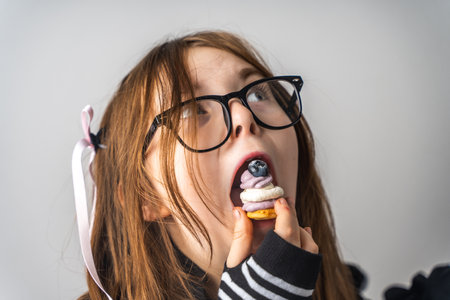 A little girl, a pre-teenager holding a cake in her hands and looking at it smiling, she is dressed in black and white clothes and black eye glasses on a gray background. High quality photoの写真素材