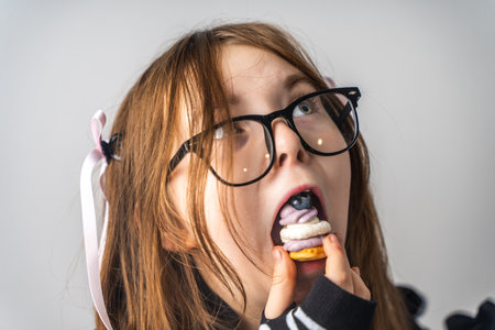 A pretty little girl, a pre-teenager holding a cake in her hands and looking at it smiling, she is dressed in black and white clothes and black eye glasses on a gray background. High quality photoの写真素材