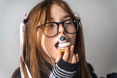 A pretty little girl, a pre-teenager holding a cake in her hands and looking at it smiling, she is dressed in black and white clothes and black eye glasses on a gray background. High quality photoの写真素材