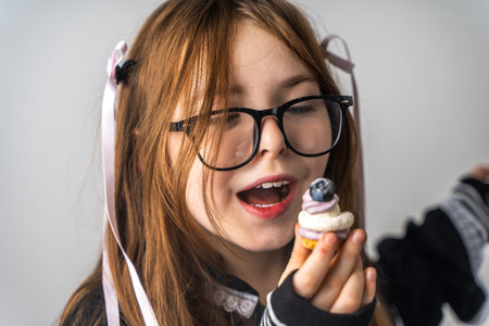 A little girl, a pre-teenager holding a cake in her hands and looking at it smiling, she is dressed in black and white clothes and black eye glasses on a gray background. High quality photoの写真素材