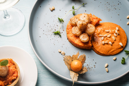 A gourmet scallops lunch with carrot puree in Romesco sauce served on a blue plate with fesalis, a glass of white wine and cutlery. High quality photoの写真素材