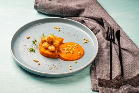 Scallops with carrot puree with Romesco sauce, fesalis and pine nuts on a blue plate on a light background with a napkin and cutlery. Diet food, vegetarian dish. High quality photoの写真素材