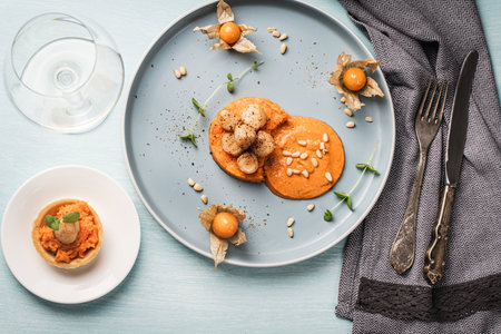 Scallops with carrot puree with Romesco sauce, fesalis and pine nuts on a blue plate on a light background with a napkin and cutlery and a glass of white wine. High quality photoの写真素材