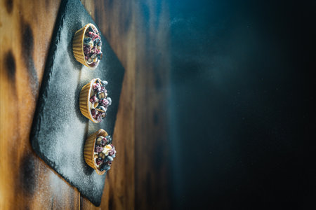 Tartlets with cottage cheese cream, cranberries and blueberries on a black plate, sprinkled with powdered sugar on a wooden background. High quality photoの写真素材