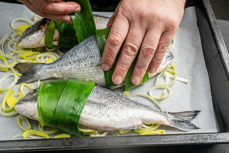 The chef's hands wrap the dorado fish in leek or palm leaves on baking paper or parchment with chopped onions on the baking tray. High quality photoの写真素材