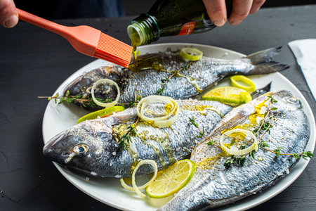Recipe for dorado fish. Raw, fresh fish with coarse salt, leeks and limes are poured with olive oil, greasing the fish with a cooking brush on a black background. High quality photoの写真素材