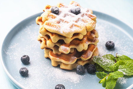 Belgian waffles stacked with blueberries, sprinkled with powdered sugar on a blue plate and mint background. High quality photoの写真素材