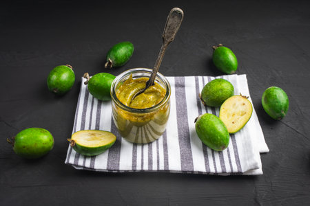 Mashed raw feijoa jam in a glass jar on a white napkin with a spoon and feijoa fruit, a healthy subtropical plant full of vitamin C, grown mainly in Latin America. Black background or table. High quality photoの写真素材