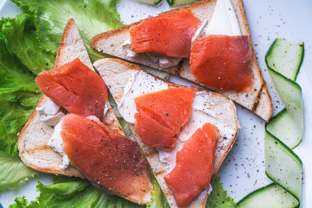 Sandwiches with cottage cheese and slices of salted trout on a white plate with cucumber and herbs....