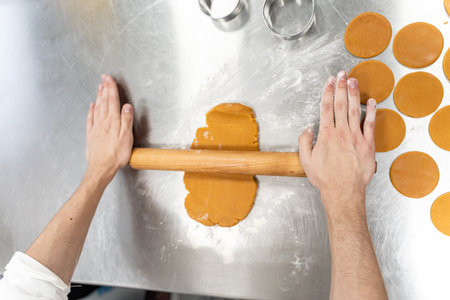 The baker's hands roll out the cookie dough on the production table with a rolling pin. The process...