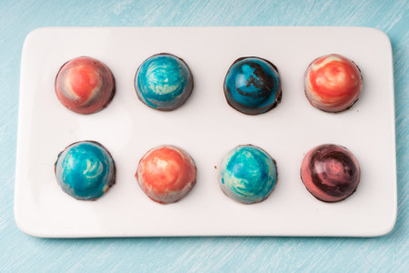 Assorted multi-colored homemade chocolates. Modern hand-painted chocolates on a white plate on a blue background. Product concept for chocolatier. Selected focus. High quality photoの写真素材