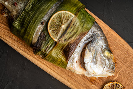 Baked Dorado fish in leek leaves with lime and lemon on a wooden board on a black background. Space for text, copyspace. High quality photoの写真素材