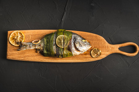 Baked Dorado fish in leek leaves with lime and lemon on a wooden board on a black background. Space for text, copyspace. High quality photoの写真素材