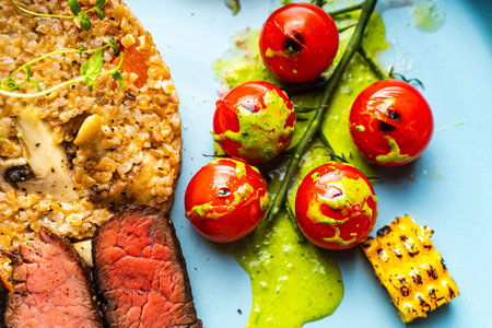Sliced filet mignon steak with grits, cherry tomatoes, mushrooms and corn is originally served on a blue plate with a napkin and cutlery on a wooden background. Top view. High quality photoの写真素材