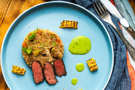 Sliced filet mignon steak with grits, mushrooms and corn served on a blue plate with a napkin and...