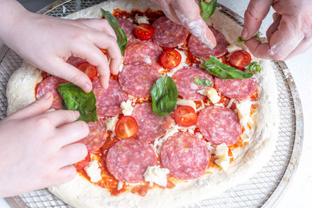 The hands of a man and a child make a paperoni pizza, spreading basil on the dough with sausage, tomatoes and cheese. The concept of family pastime, hobbies. High quality photoの写真素材