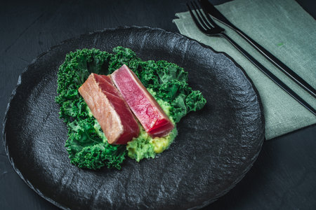 The tuna slices are lightly fried on mashed potatoes with spinach and Kale lettuce on a black mica plate and background. The concept of healthy eating. Original serving of the dish. High quality photoの写真素材