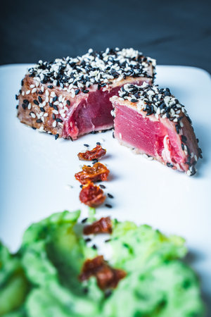 Tuna slices with green puree, sesame seeds and sun-dried tomatoes on a white plate on a dark background. Restaurant serving fish. Healthy eating. Vertical photo. High quality photoの写真素材