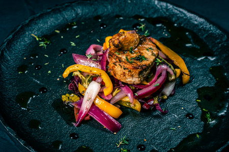Slice of roasted pork tenderloin with vegetables, pink onion and yellow pepper on a black plate on a dark background. Beautiful, restaurant presentation of the dish. High quality photoの写真素材