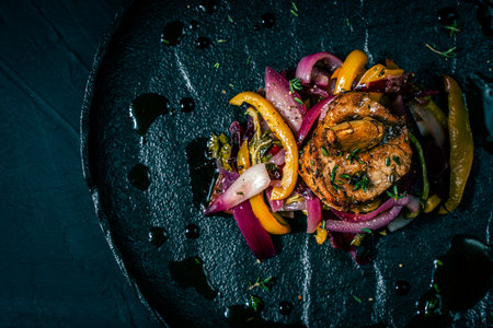 Slice of roasted pork tenderloin with vegetables, pink onion and yellow pepper on a black plate on a dark background. Beautiful, restaurant presentation of the dish. Copy space. High quality photoの写真素材