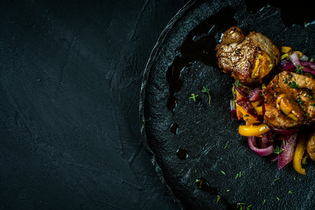 Slice of roasted pork tenderloin with vegetables, pink onion and yellow pepper on a black plate on a dark background. Beautiful, restaurant presentation of the dish. Copy space. High quality photoの写真素材