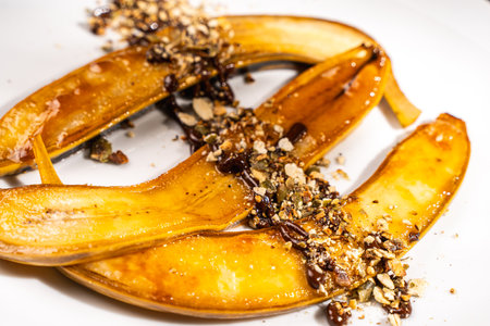 Flambe bananas. Dessert of bananas with pistachios, toasted pumpkin seeds, chocolate and caramel on a white plate on a dark green background. Place for text. Top view. Close-up. High quality photoの写真素材