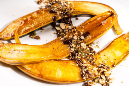 Flambe bananas. Dessert of bananas with pistachios, toasted pumpkin seeds, chocolate and caramel on a white plate on a dark green background. Place for text. Top view. Close-up. High quality photoの写真素材