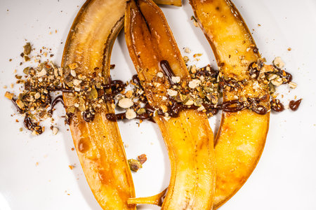 Flambe bananas. Dessert of bananas with pistachios, toasted pumpkin seeds, chocolate and caramel on a white plate on a dark green background. Place for text. Top view. Close-up. High quality photoの写真素材
