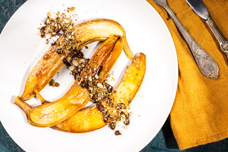 Flambe bananas. Dessert of bananas with pistachios, toasted pumpkin seeds, chocolate and caramel on a white plate on a dark green background. Place for text. Top view. Close-up. High quality photoの写真素材