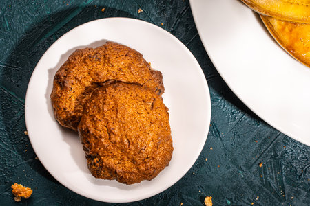 Close-up of two homemade cookies on a white plate, with a side plate of caramelized bananas with pistachios, toasted pumpkin seeds, chocolate, and caramel on a dark green background. Place for text. High quality photoの写真素材