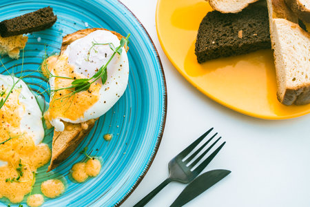Poached eggs with hollandaise sauce on toast with crouton on a turquoise plate, next to a glass of orange juice and cutlery. High quality photoの写真素材
